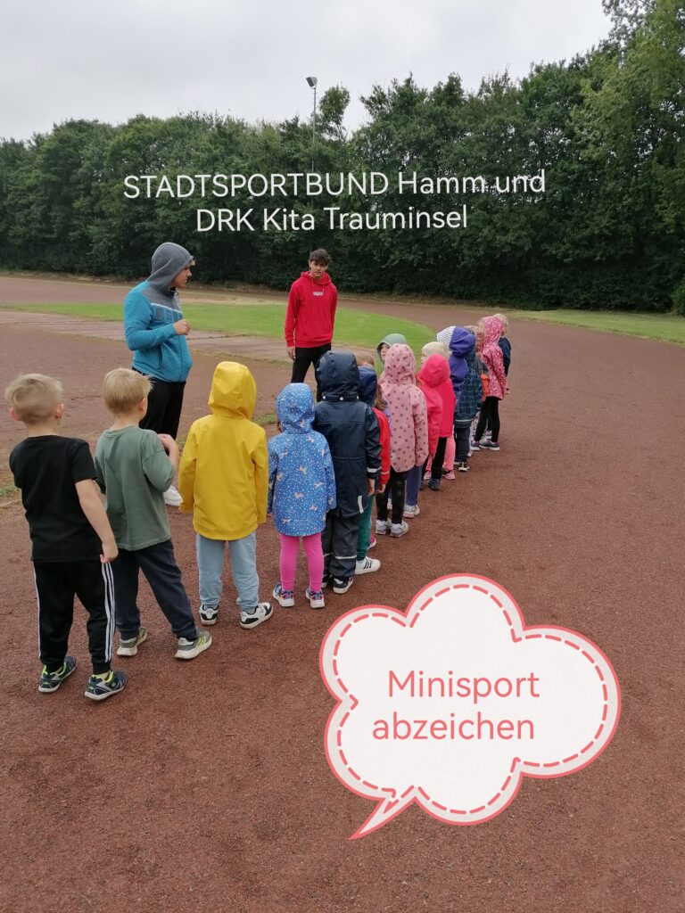 Mehrere Kinder stehen mit bunten Regenjacken auf einem Sportplatz. Sie stehen mit dem Rücken zum Fotografen. Vor den Kindern stehen zwei junge Männer. Einer in einem roten Pullover, einer in einem blau, grauen. 
